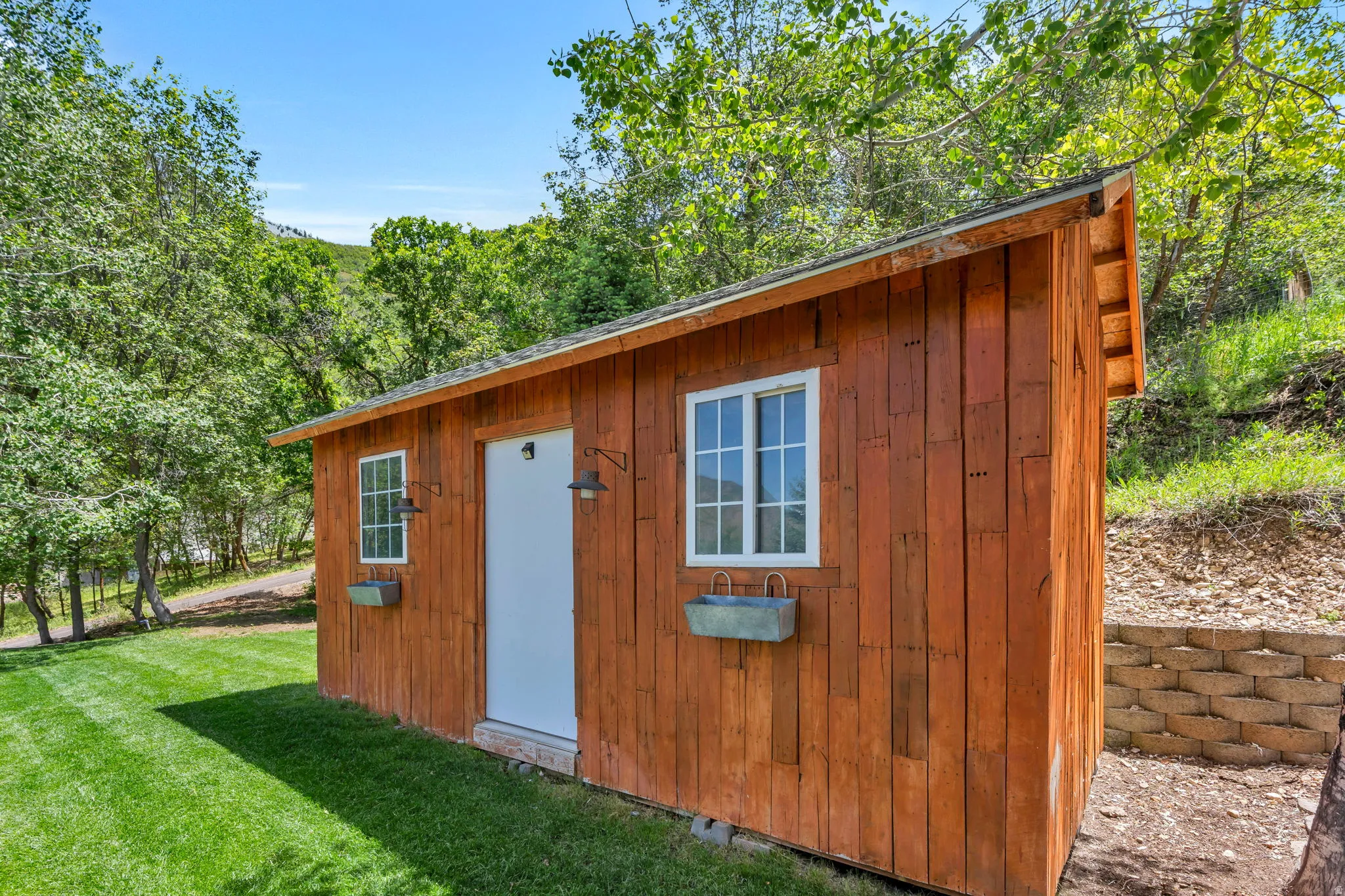Wood-sided outbuilding featuring a white entry door, two multi-pane windows, and exterior light fixtures