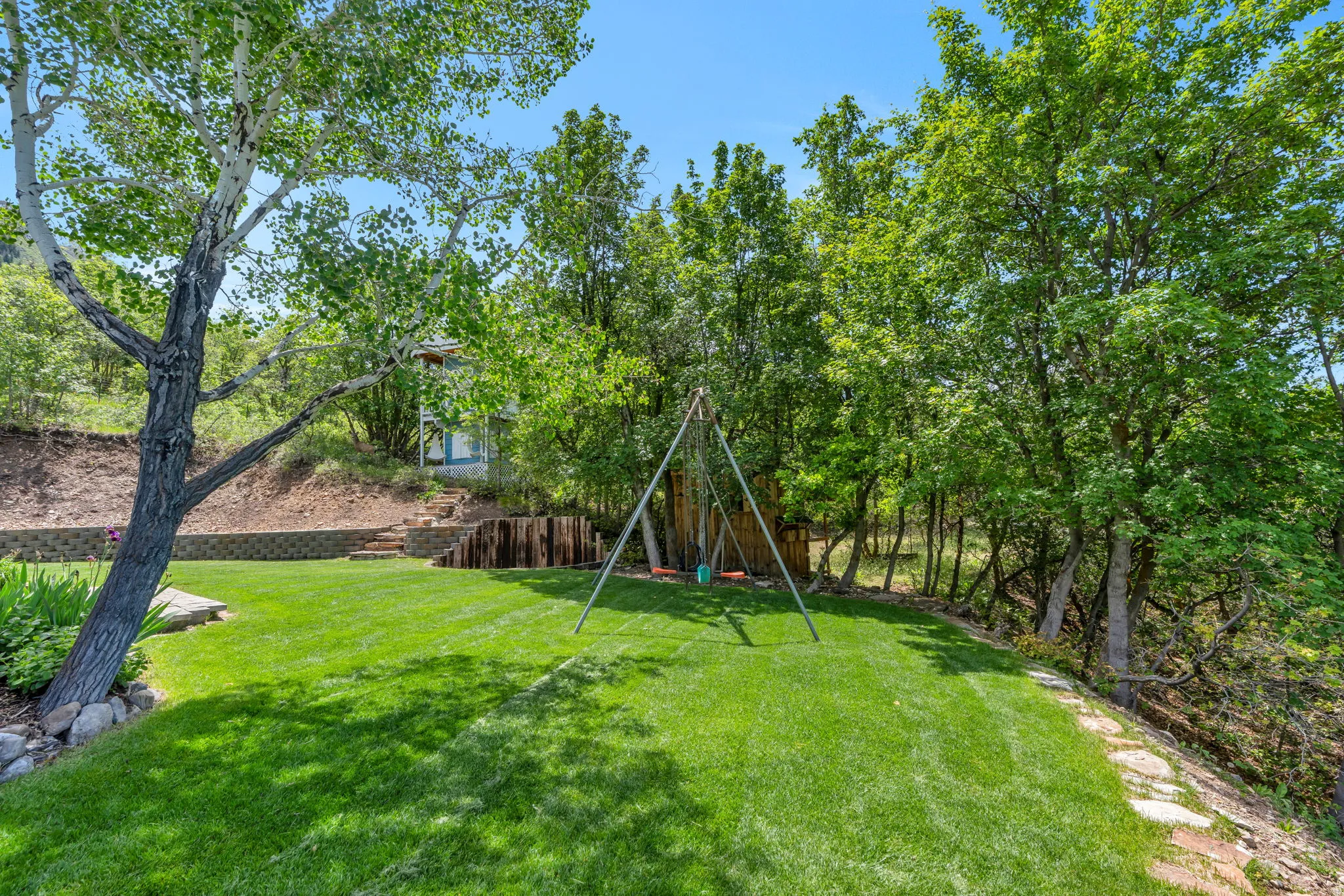 Expansive sloped lawn area featuring mature trees, a stone retaining wall, and natural wood steps