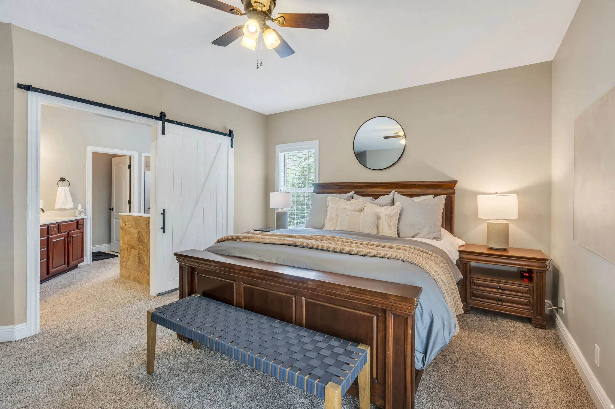 Carpeted sleeping area featuring a ceiling fan with integrated lighting, a window with natural light, and a sliding barn door entry to a bathroom with a vanity and sink
