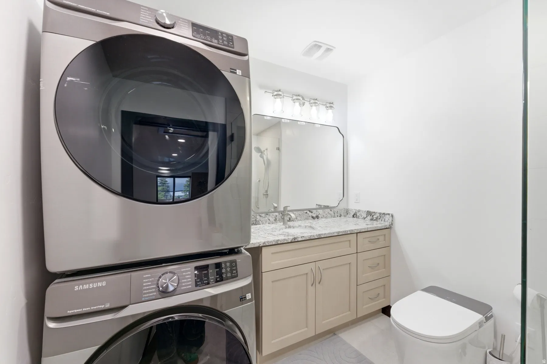 Integrated laundry appliances, light wood-finish vanity with drawer storage, granite countertop, undermount sink with a chrome faucet, wall-mounted mirror, and glass shower partition