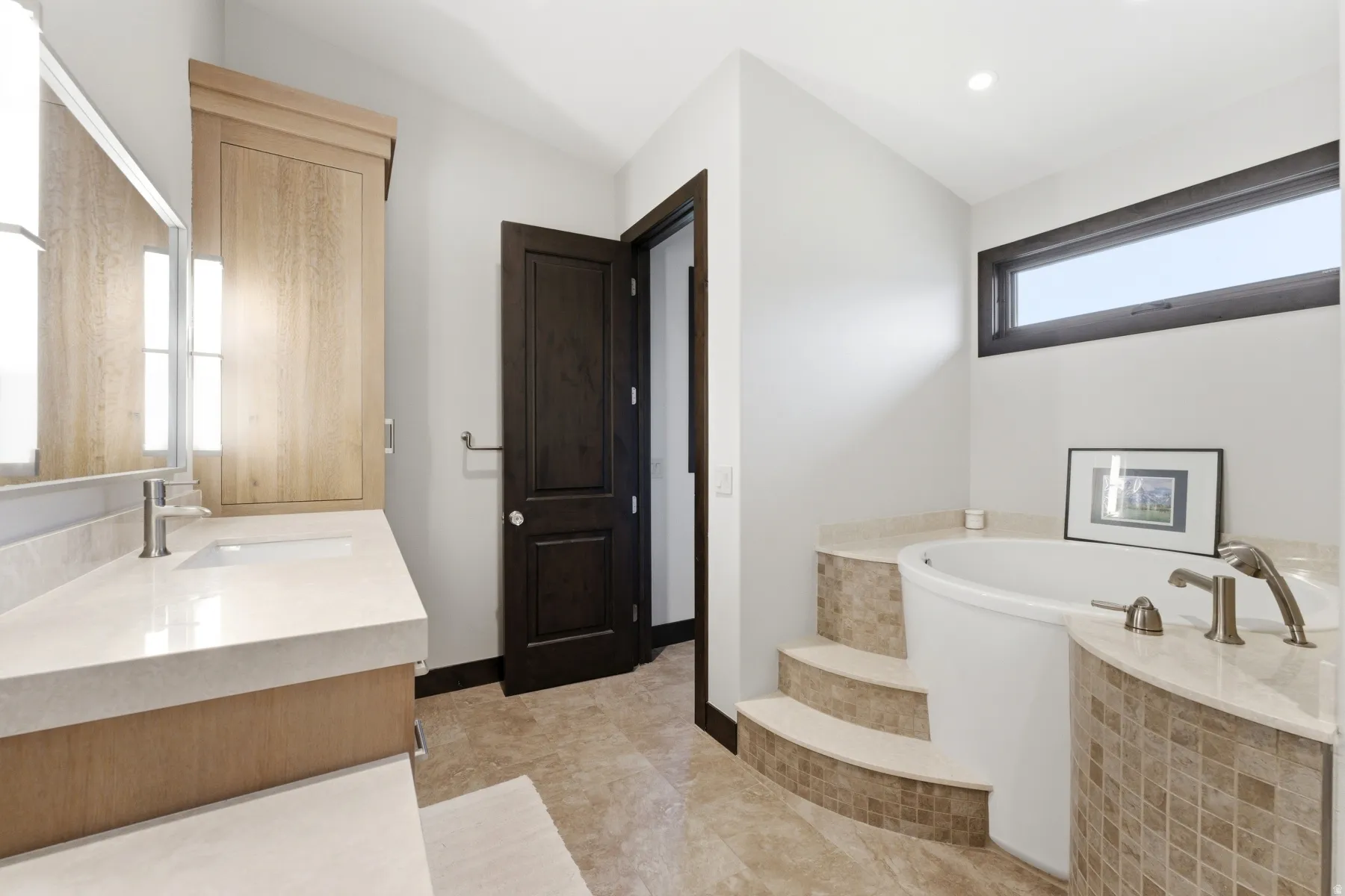 Contemporary bathroom featuring a built-in soaking tub with mosaic tile surround, a light wood-finish vanity with an integrated sink, a large mirror with integrated lighting, and tile flooring