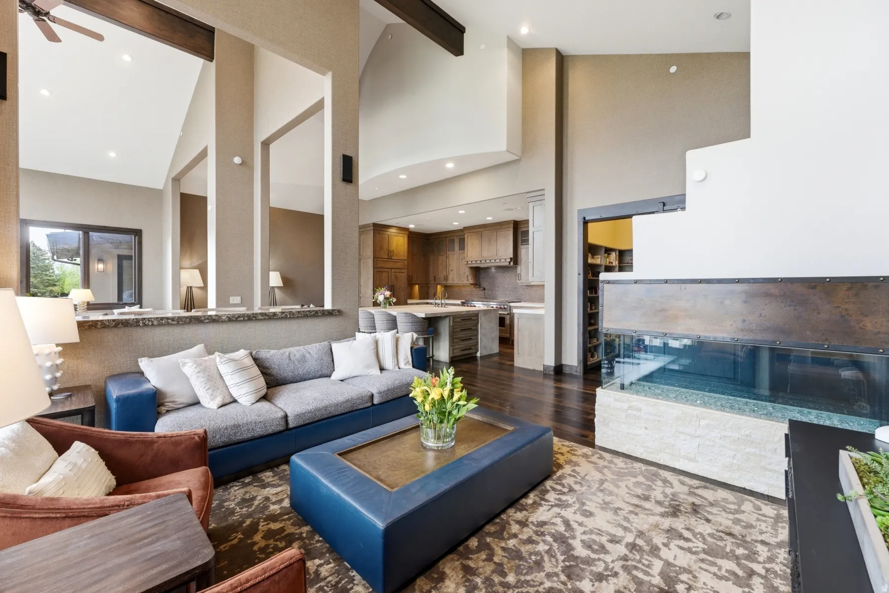 Open-concept living area featuring a vaulted ceiling with exposed beams, a gas fireplace with a glass enclosure and stacked stone base, wood-finish flooring, and integrated ceiling speakers