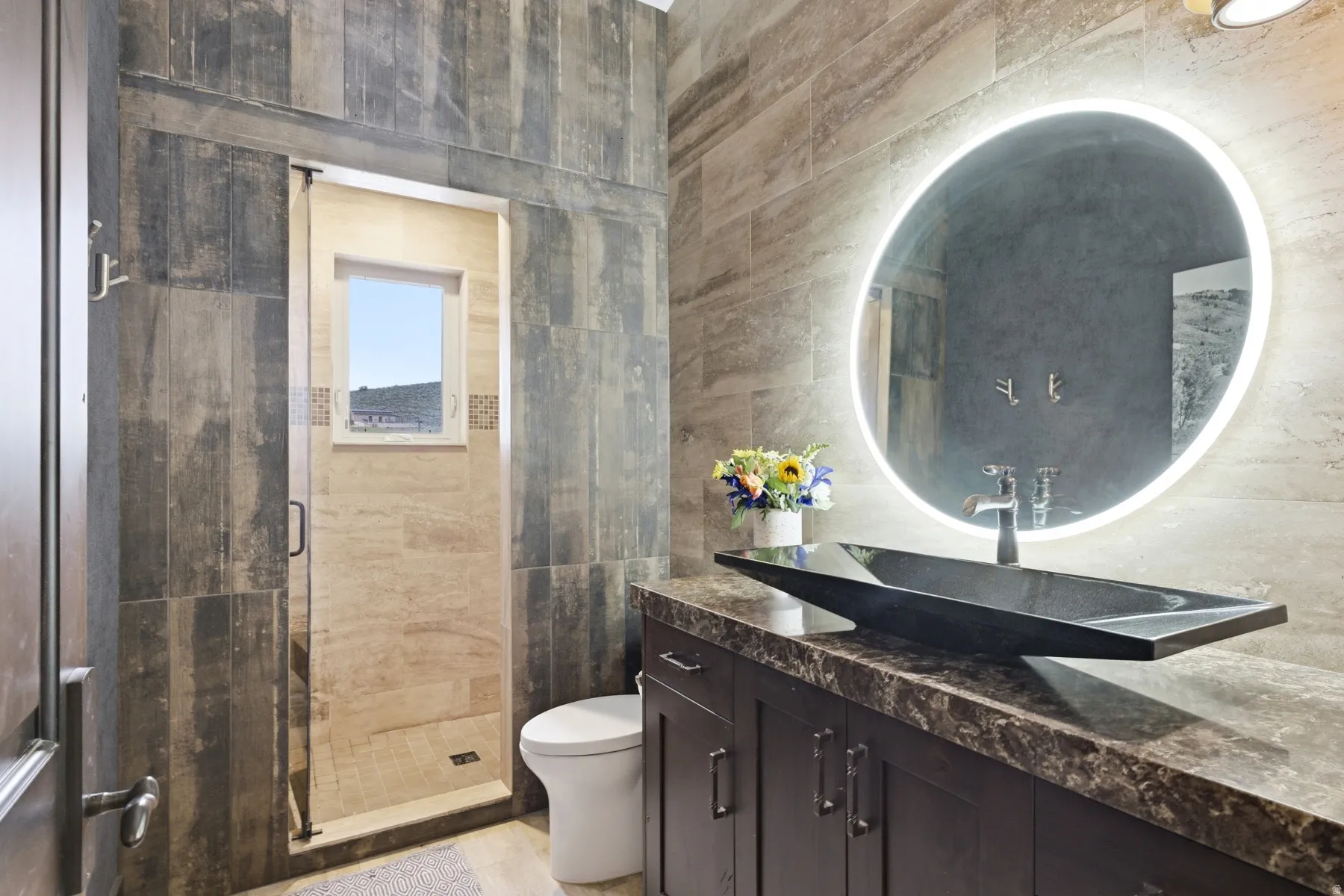 Bathroom featuring a contemporary vanity with an integrated vessel sink, an illuminated mirror, and a walk-in shower with natural stone-look tiling