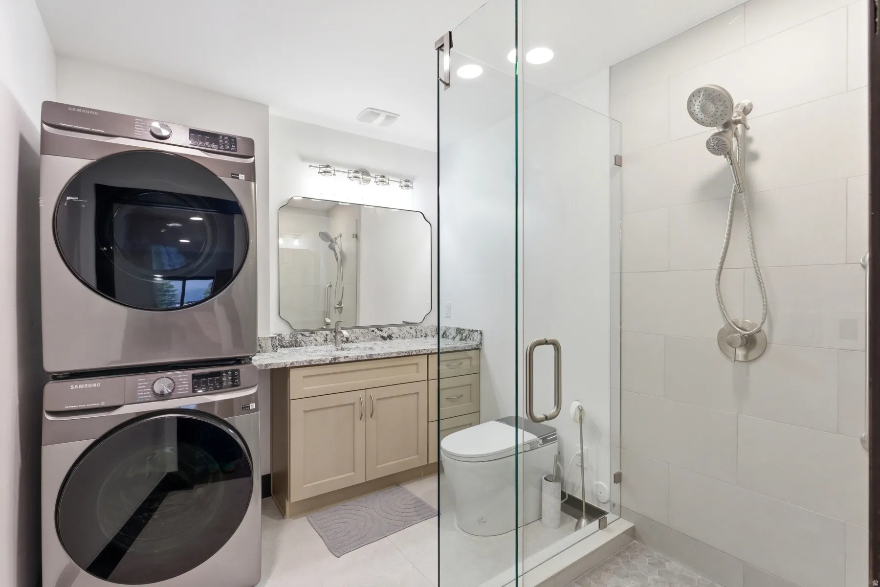 Modern bathroom featuring a stacked laundry unit, a glass-enclosed shower with a handheld fixture, light wood-finish vanity with a stone countertop, and a decorative wall mirror