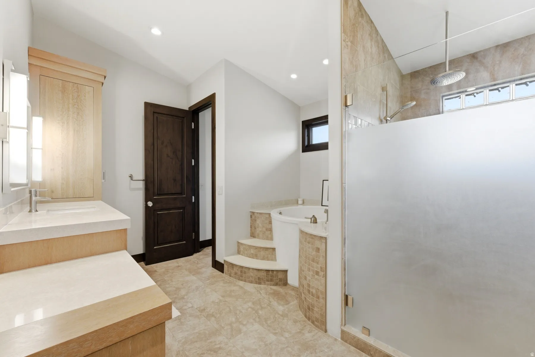 Bathroom featuring a step-up jetted tub with mosaic tile surround, a frosted glass shower enclosure with a rainfall showerhead, and a vanity with dual sinks and light-toned countertops