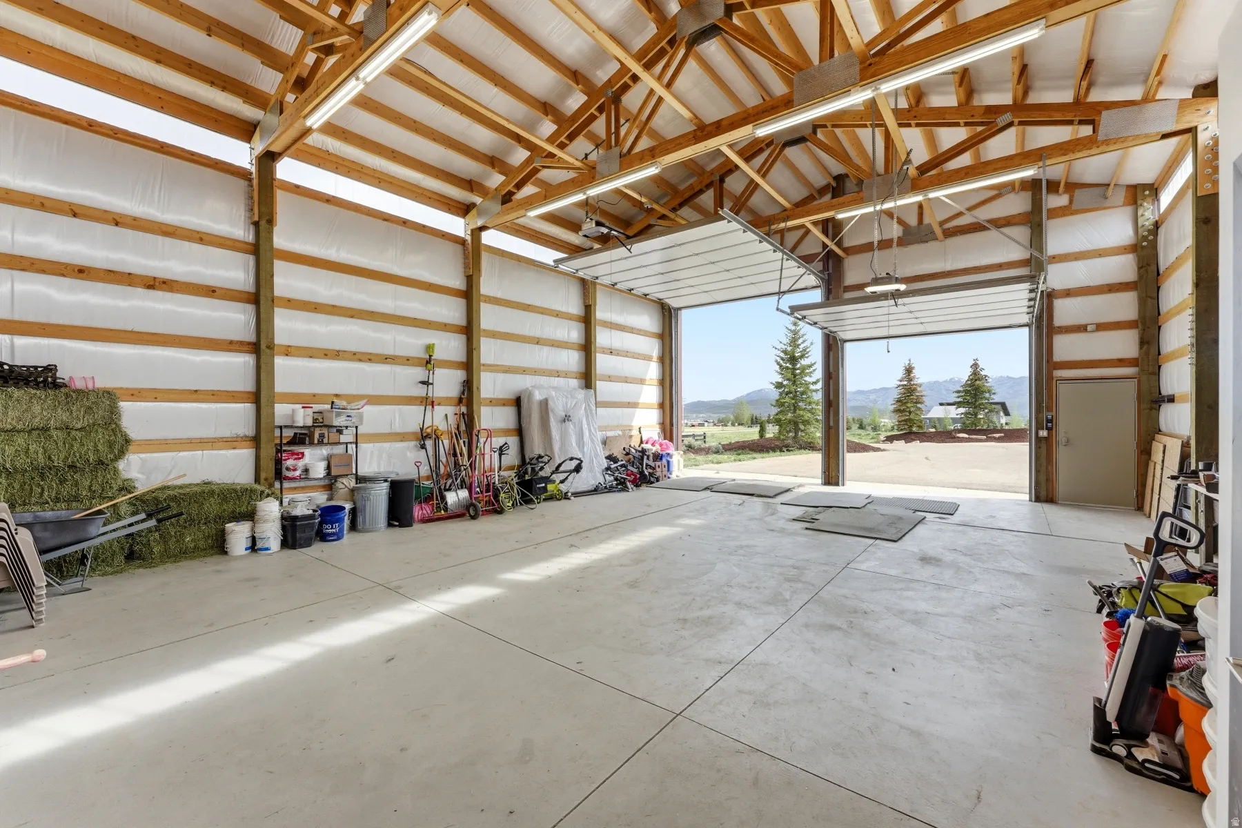 Spacious open-span structure featuring two overhead doors, concrete flooring, exposed wood beam ceilings, and fluorescent lighting