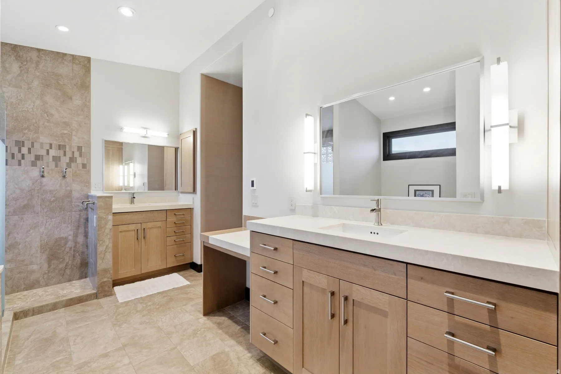 Spacious bathroom featuring light wood-finish cabinetry, dual vanity areas with undermount sinks, and stone-look tile flooring