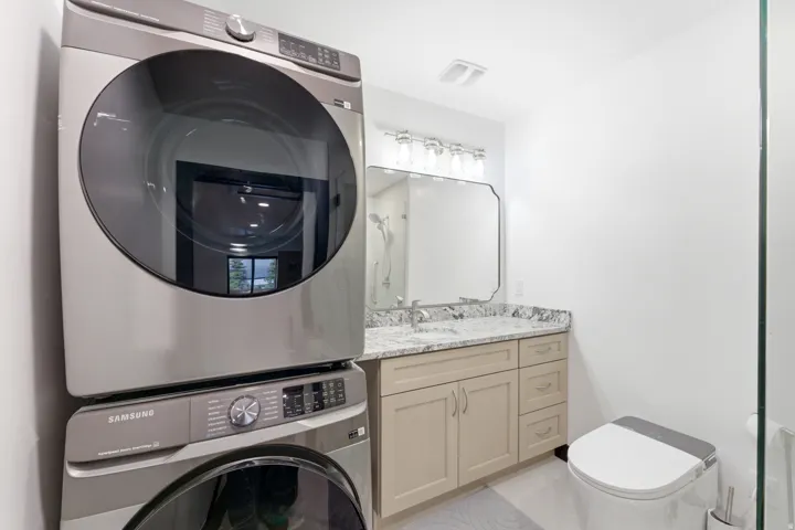 Integrated laundry appliances, light wood-finish vanity with drawer storage, granite countertop, undermount sink with a chrome faucet, wall-mounted mirror, and glass shower partition