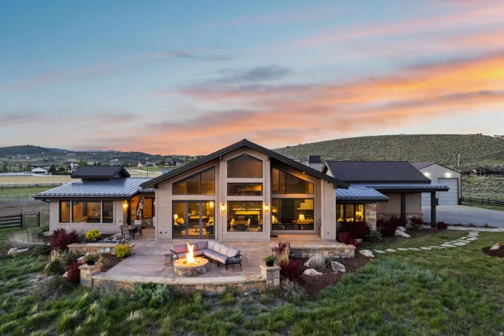 Contemporary-style residence featuring a vaulted ceiling, extensive window walls, a stone-clad patio with a built-in fire pit, metal roofing, and a detached garage structure