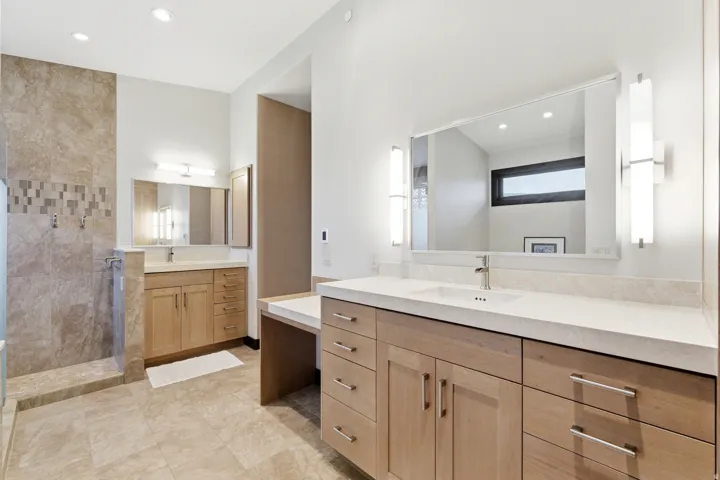 Spacious bathroom featuring light wood-finish cabinetry, dual vanity areas with undermount sinks, and stone-look tile flooring