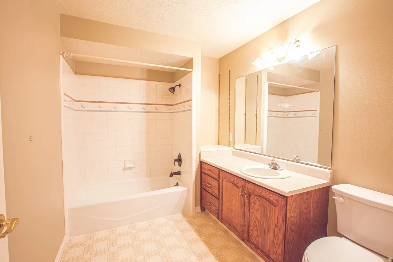 Bathroom featuring a built-in tub and shower combination with white tile surround, a vanity with a wood-finish cabinet and integrated sink, a large wall-mounted mirror, and patterned resilient flooring