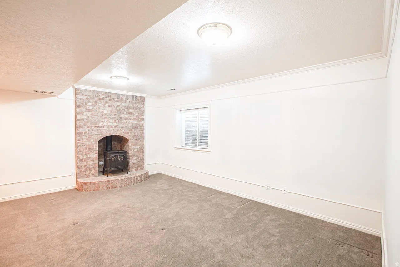 Spacious room featuring a brick fireplace with a built-in stove, neutral carpet flooring, a window with horizontal blinds, white walls, and ceiling-mounted lighting fixtures