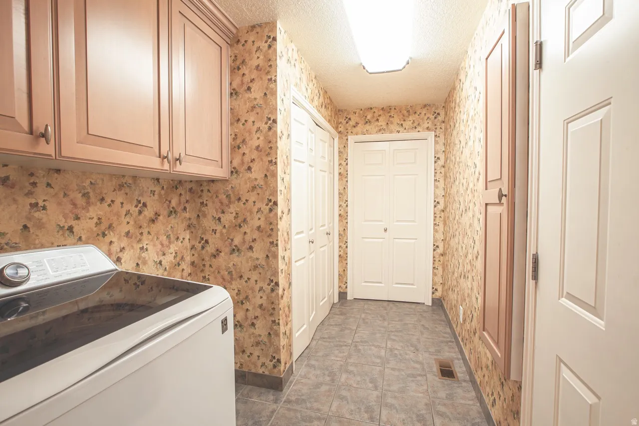 Laundry area featuring overhead cabinetry, patterned wall covering, tile flooring, and two bifold closet doors