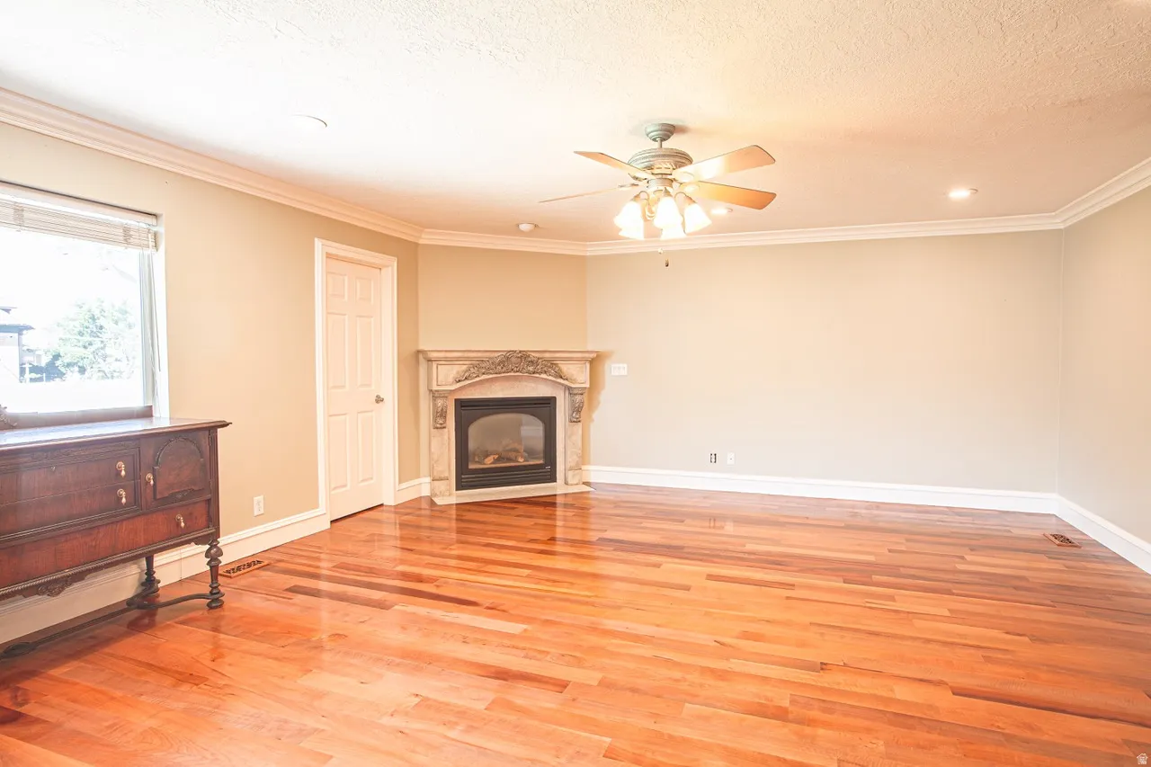 Spacious interior featuring wood-finish flooring, an ornate fireplace, crown molding, a ceiling fan with integrated lighting, and a large window