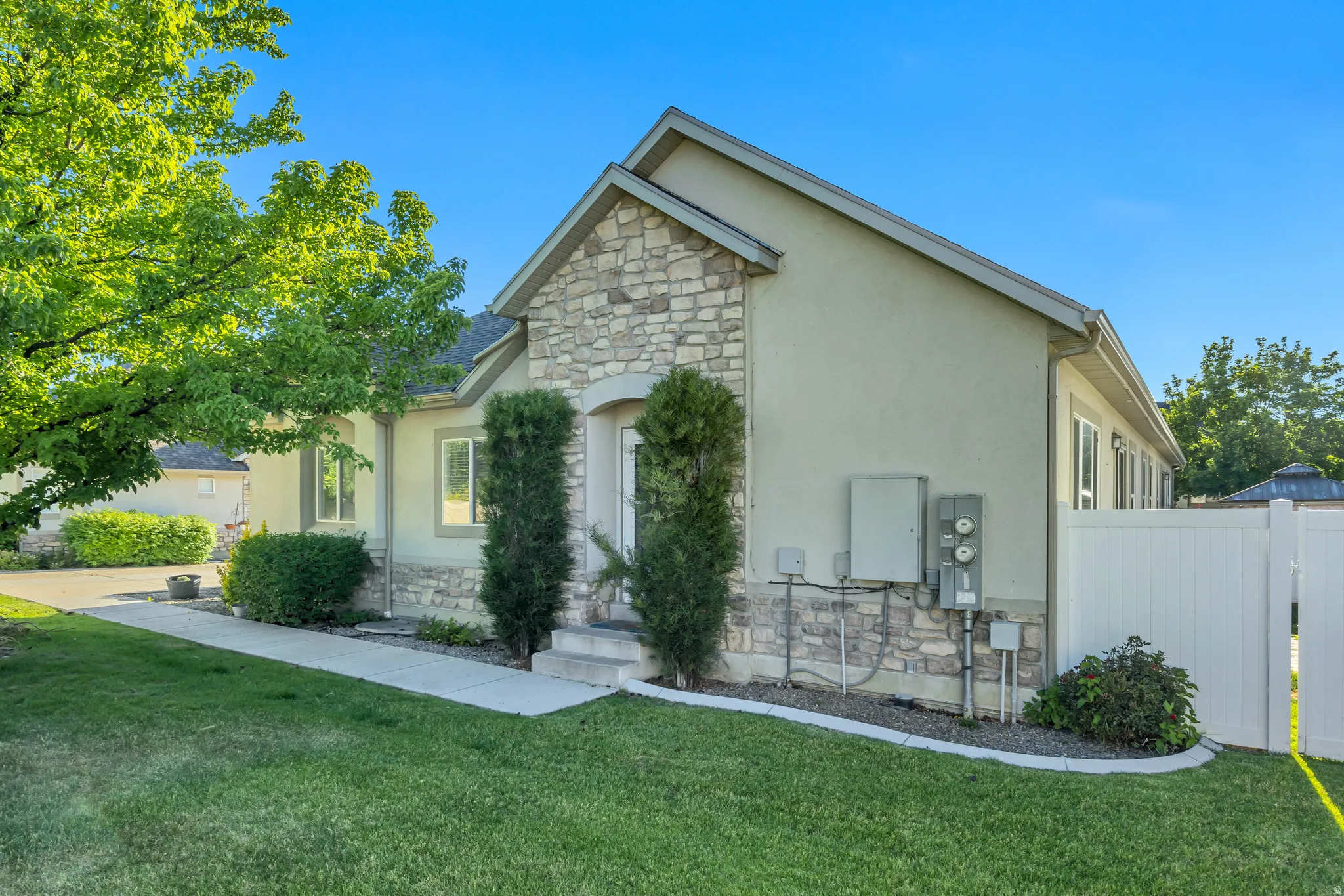 Stucco exterior with stone accents, a concrete walkway, mature landscaping, a manicured lawn, and a white privacy fence