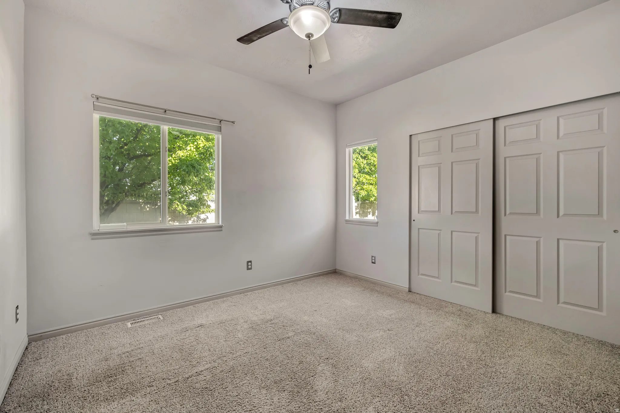 Bright room featuring two windows with natural light, neutral carpeting, a ceiling fan with integrated lighting, and two sliding closet doors