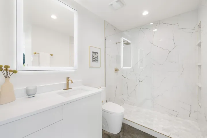 Vanity with integrated sink and gold-tone faucet, illuminated mirror, walk-in shower with full-height tile, and glass enclosure