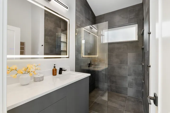 Modern bathroom featuring a single vanity with a white countertop and dark gray cabinetry, an illuminated mirror, a glass-enclosed shower with dark gray tile, and a rectangular window