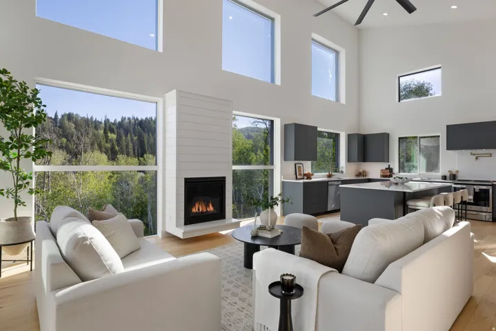 Soaring ceilings with extensive window placements, a sleek fireplace with a shiplap surround, wood-finish flooring, and a contemporary kitchen with dark cabinetry and an island