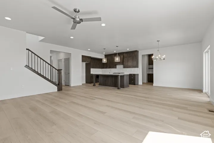 Spacious open-concept living area featuring light wood-finish flooring, a contemporary kitchen with dark wood cabinetry and a central island, a modern chandelier, and a staircase with dark wood handrails and black balusters