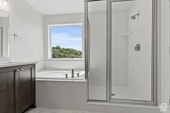 Bathroom featuring a corner soaking tub with a window, a dark wood-finish vanity with a light-toned countertop, and a glass-enclosed shower with hexagonal tile flooring and brushed nickel fixtures