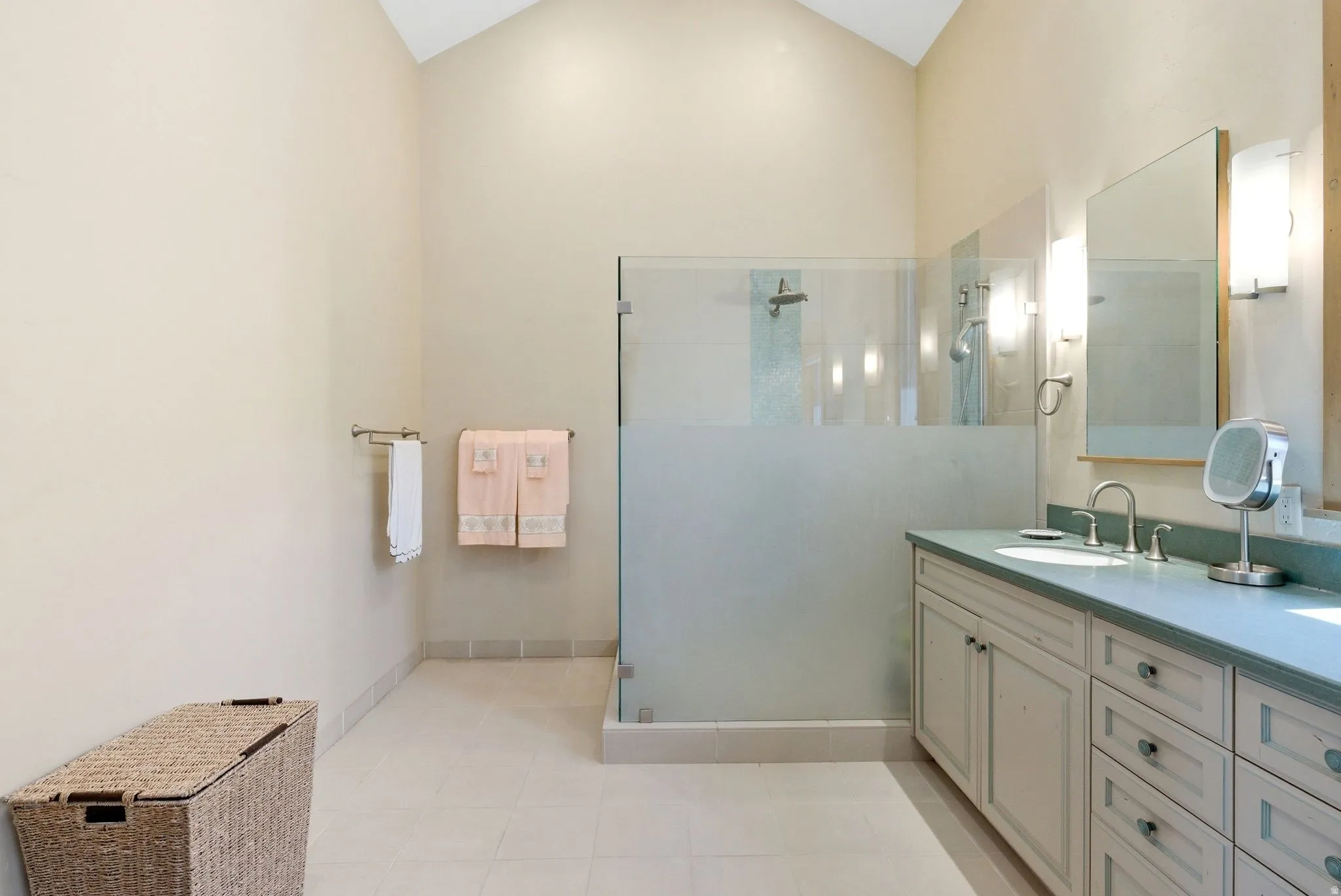 Bathroom featuring a vaulted ceiling, a spacious vanity with an undermount sink, a large mirror with sconce lighting, a glass-enclosed shower, and neutral tile flooring