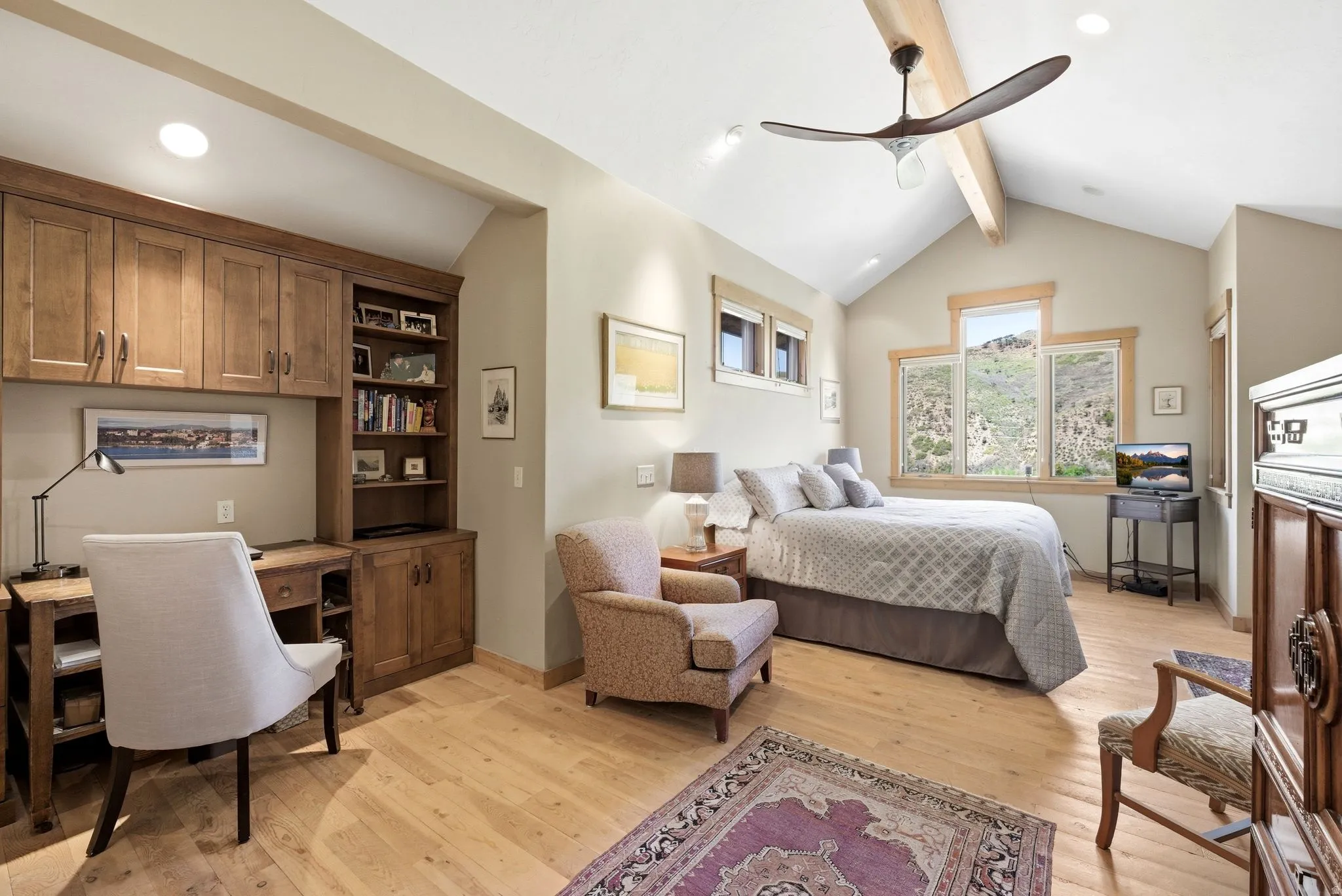 Vaulted ceiling with exposed wood beam, natural wood-finish flooring, built-in cabinetry with desk, and multiple windows offering hillside scenery