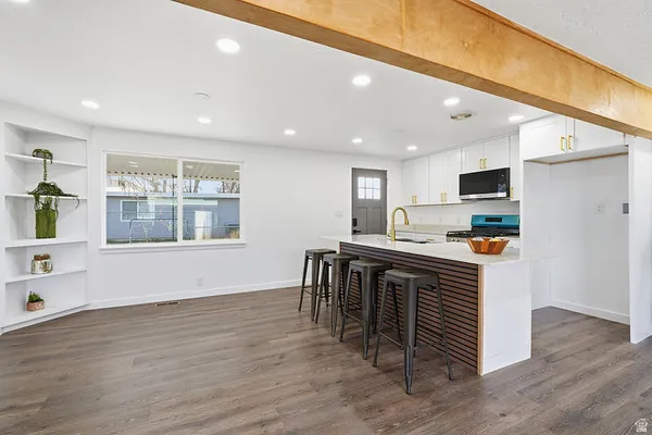 Kitchen and living area featuring wood-finish flooring, white cabinetry with gold-tone hardware, a kitchen island with seating, a window overlooking the exterior, and built-in shelving