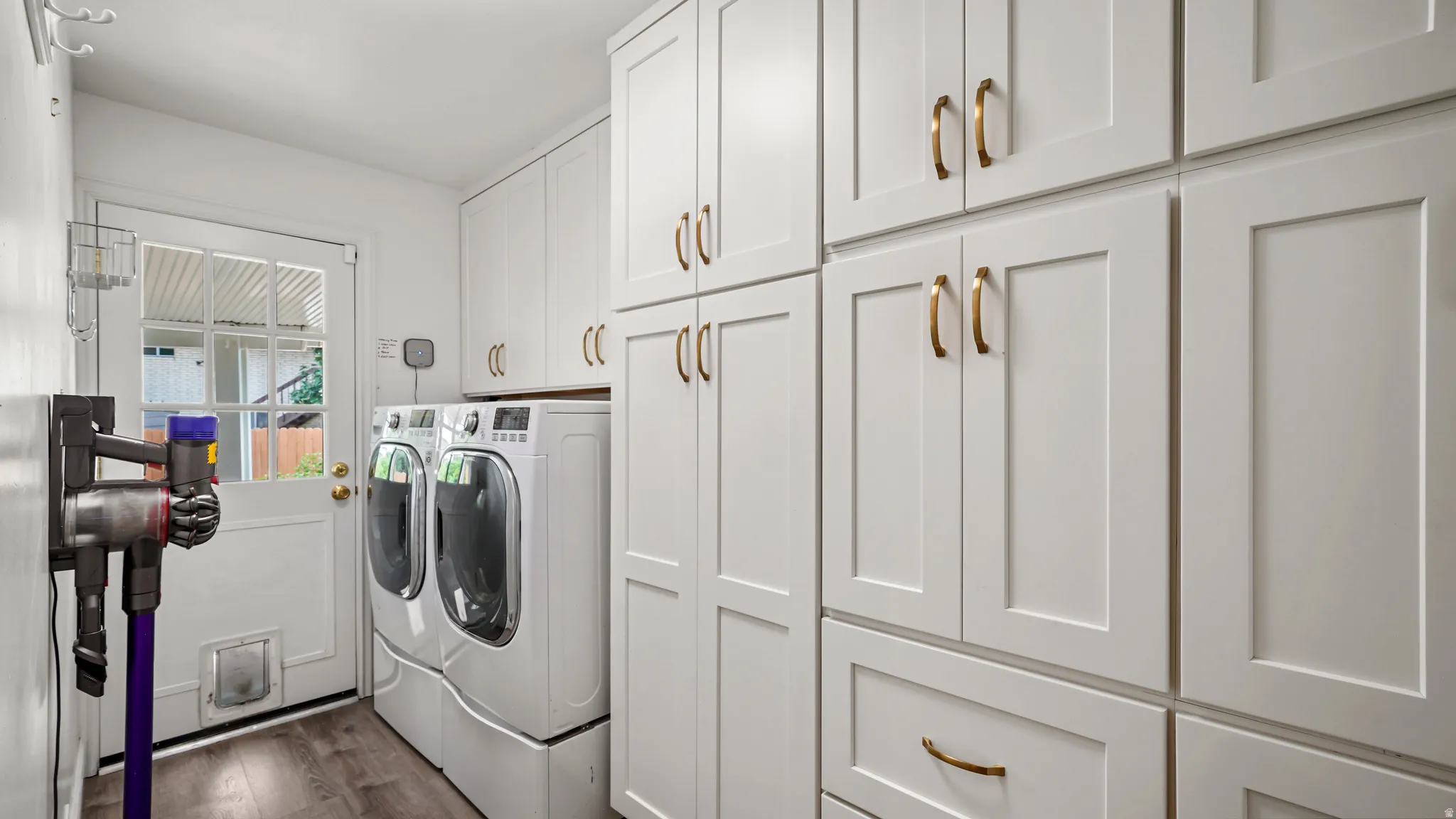 Laundry room featuring extensive white shaker cabinetry, gold-toned hardware, wood-finish flooring, and a glass-paneled door