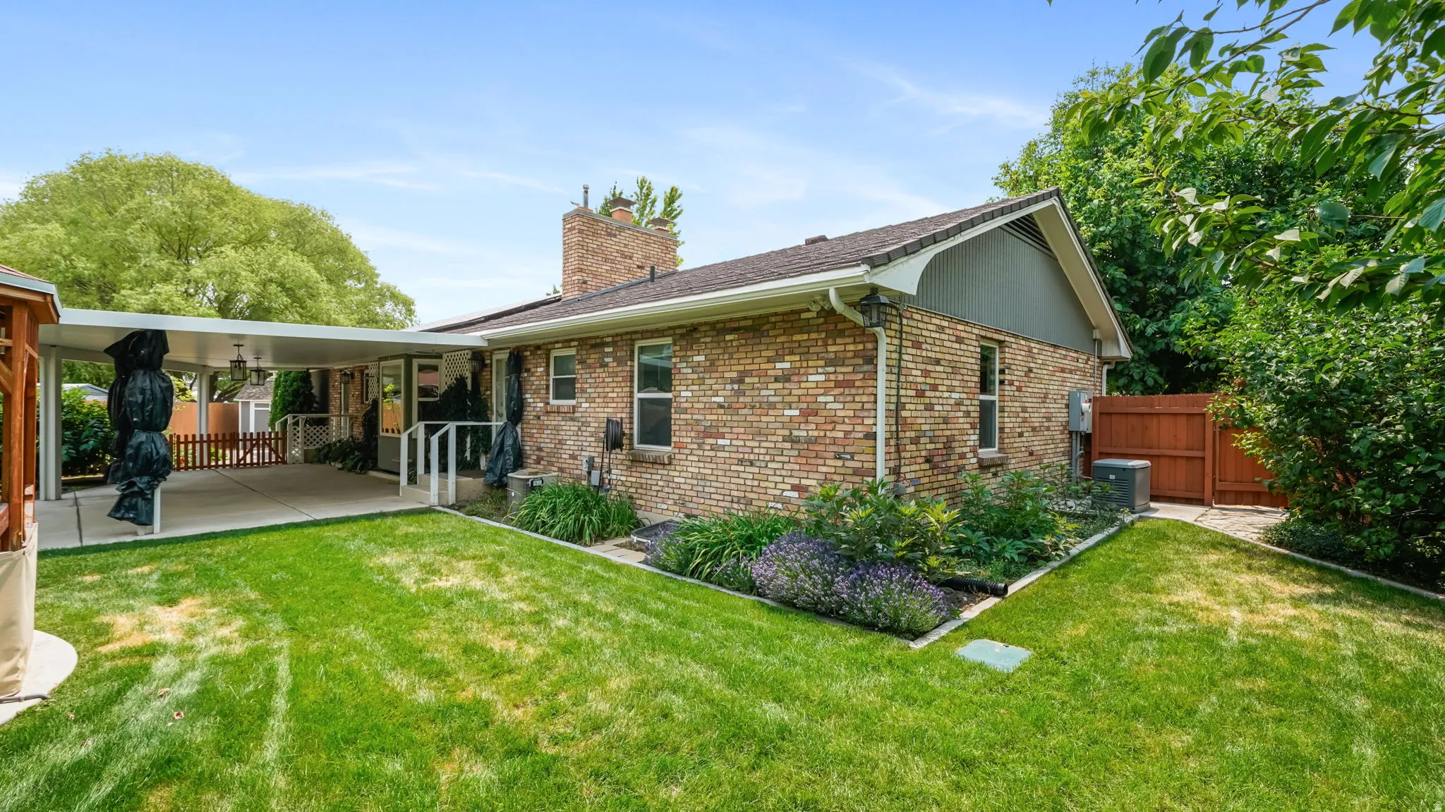 Expansive backyard featuring a manicured lawn, a covered patio, brick exterior, mature trees, and a privacy fence