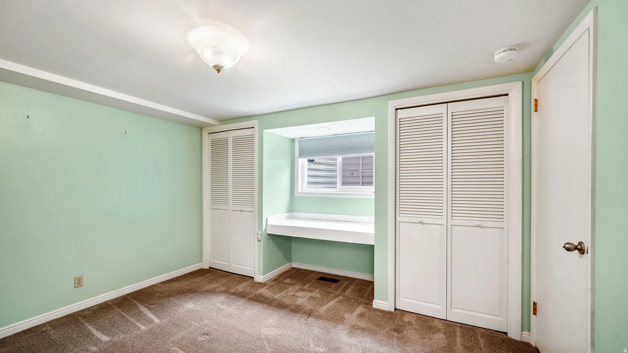Room featuring a built-in window desk, two sets of louvered bi-fold closet doors, wall-to-wall carpeting, and a flush-mount ceiling light fixture