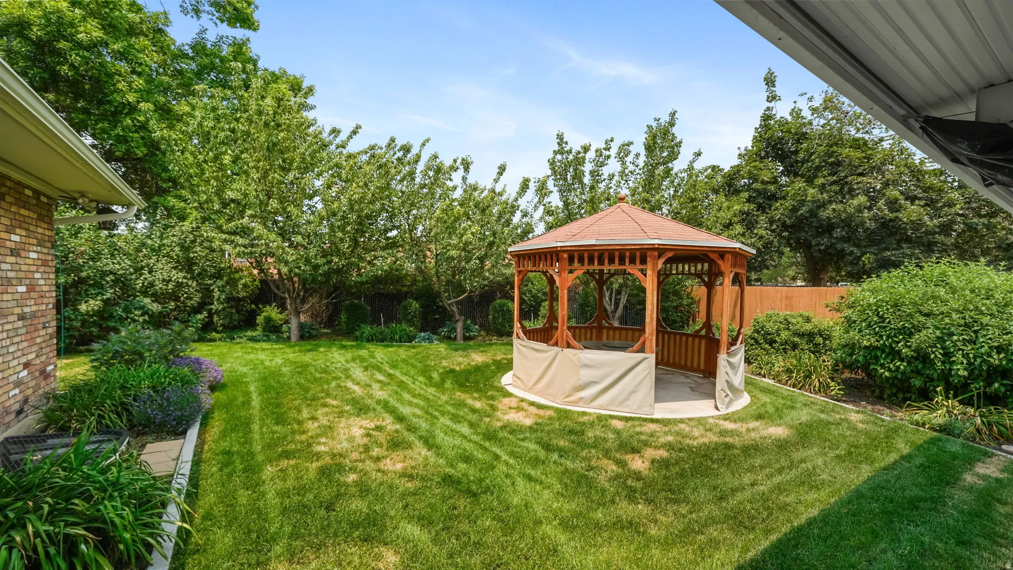 Expansive backyard featuring a central wooden gazebo with a shingled roof, a manicured lawn, established perimeter trees, and landscaping beds with various plantings