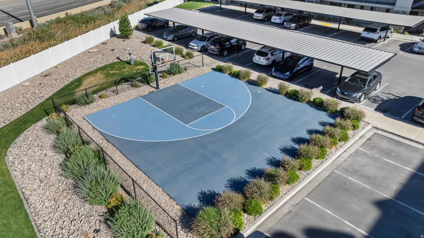 Outdoor basketball court featuring a blue playing surface with perimeter landscaping, fencing, and adjacent covered parking