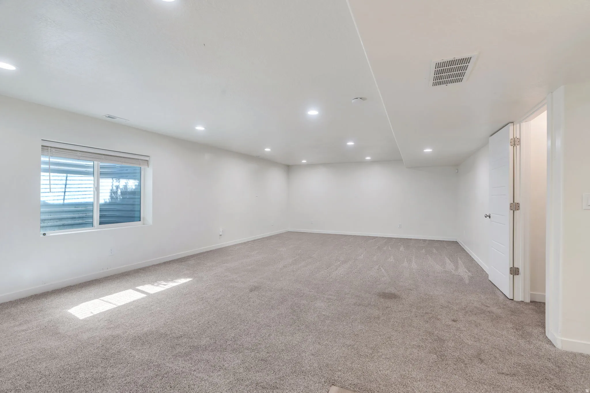 Spacious room featuring light gray carpet flooring, recessed ceiling lighting, and a window with blinds