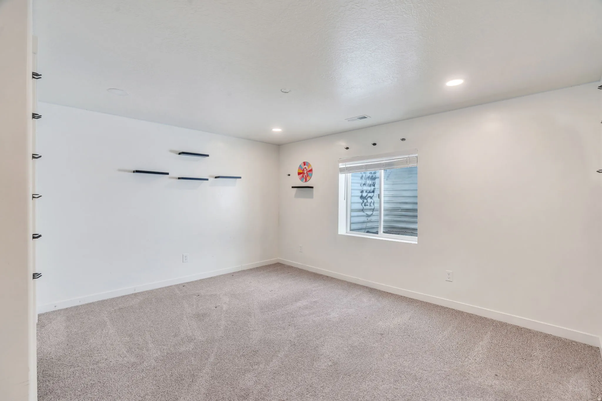 Spacious room featuring light-toned carpeting, recessed ceiling lighting, a window with blinds, and white wall finishes