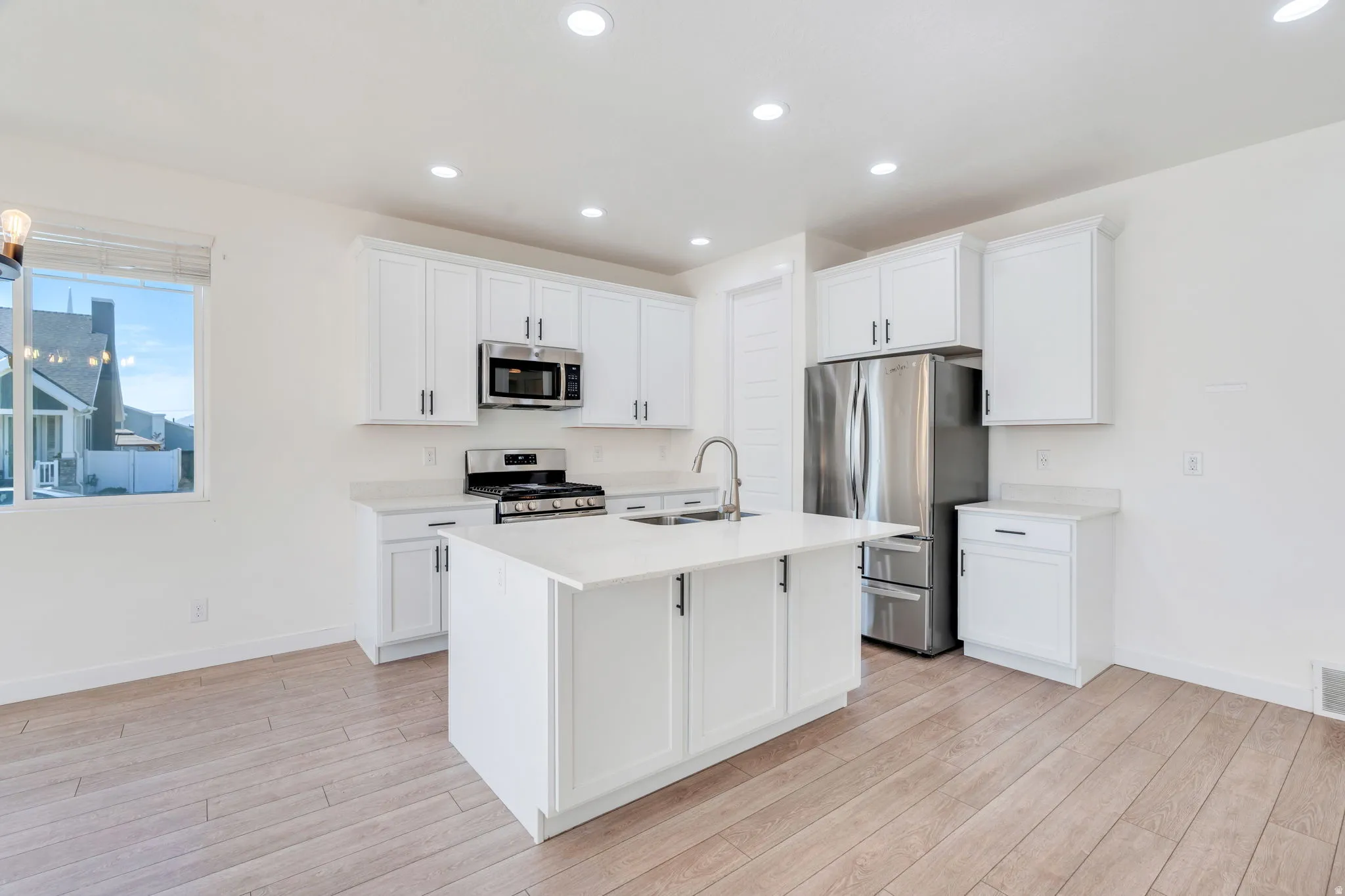 Bright kitchen featuring white shaker cabinetry, stainless steel appliances, a center island with sink, light-toned wood-finish flooring, and recessed lighting