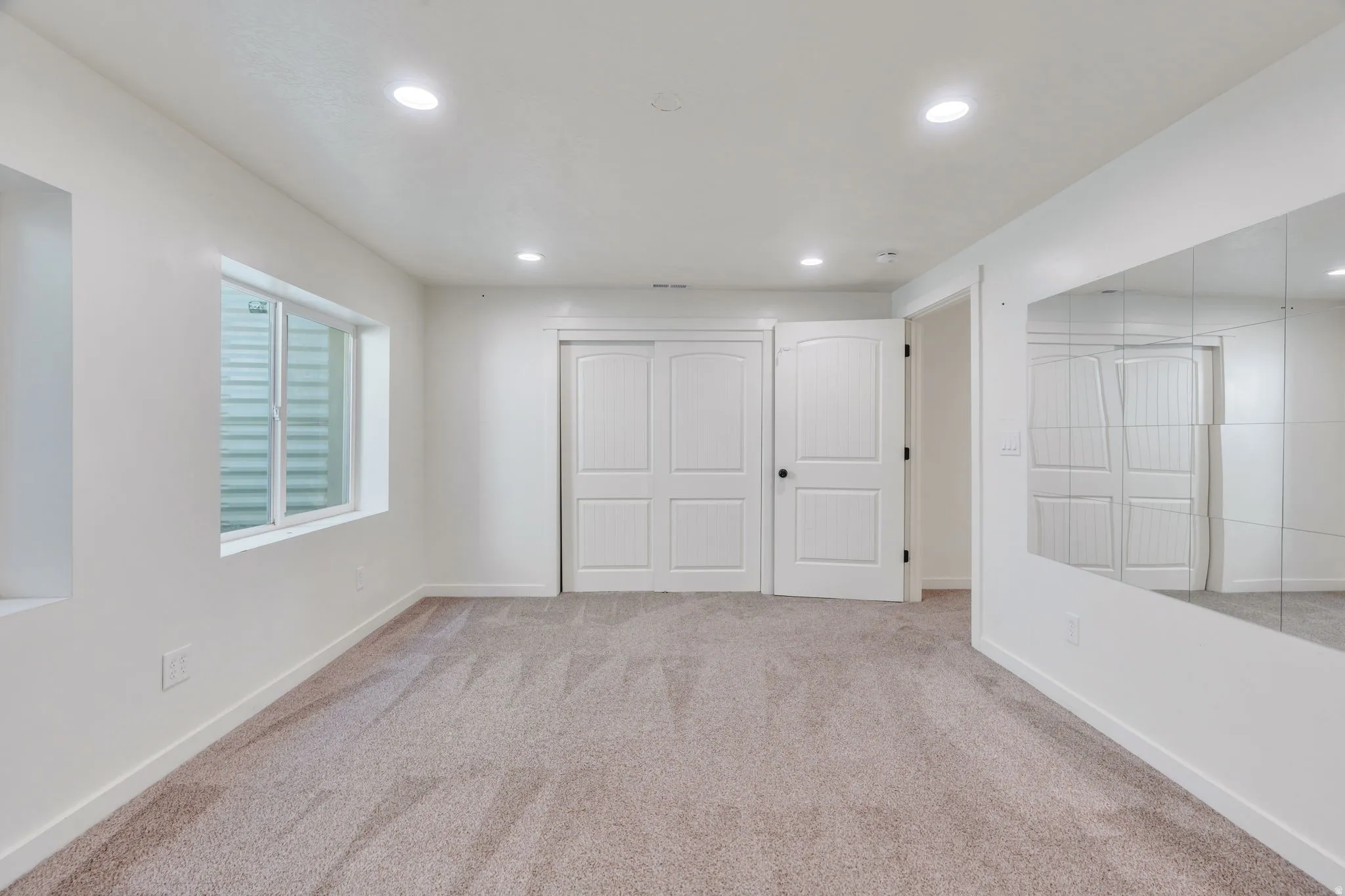 Spacious room featuring neutral carpeting, recessed lighting, a large window, white paneled doors, and a mirrored wall