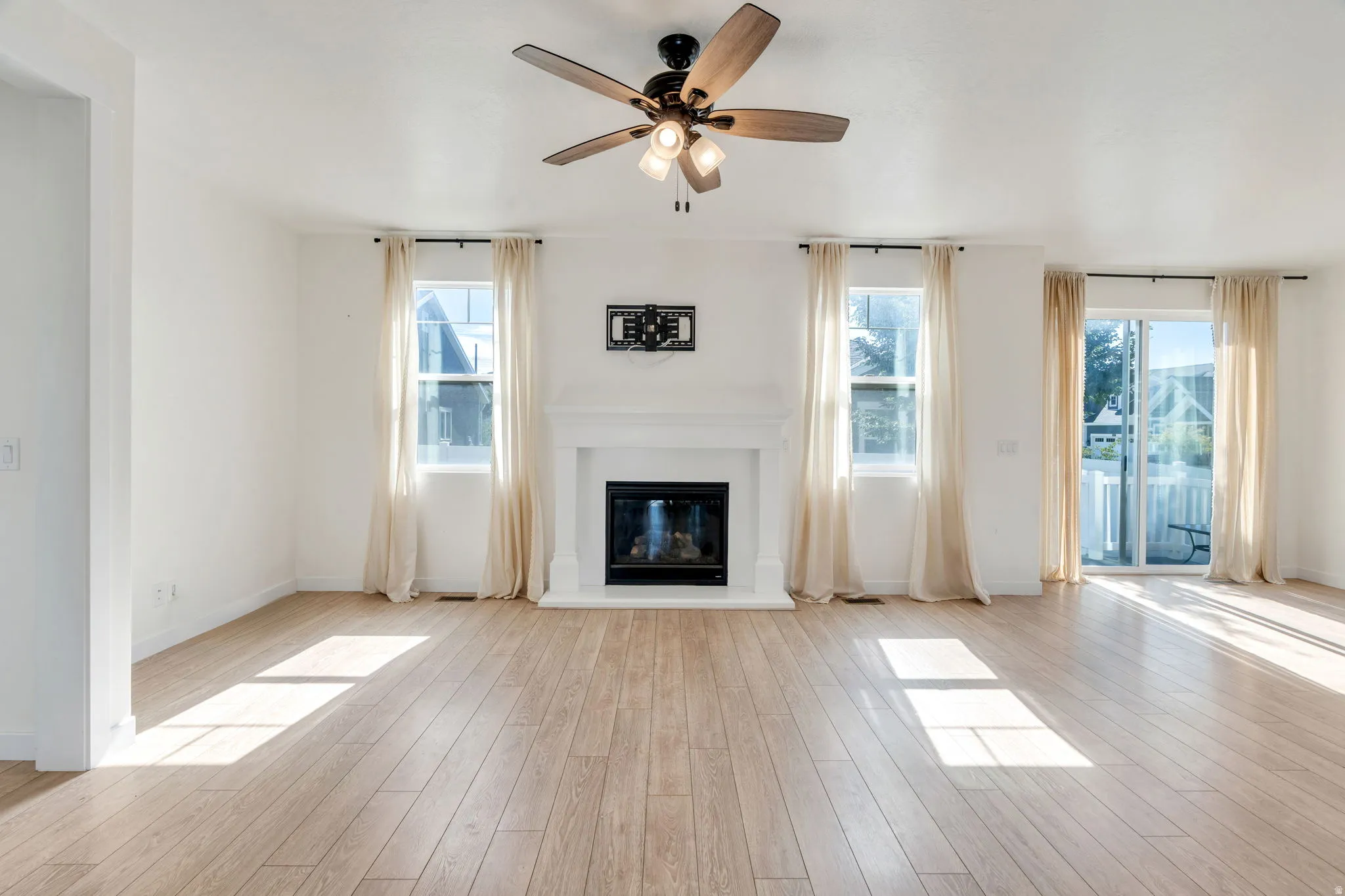 Spacious room featuring light wood-finish flooring, a ceiling fan, and a fireplace with a white mantel