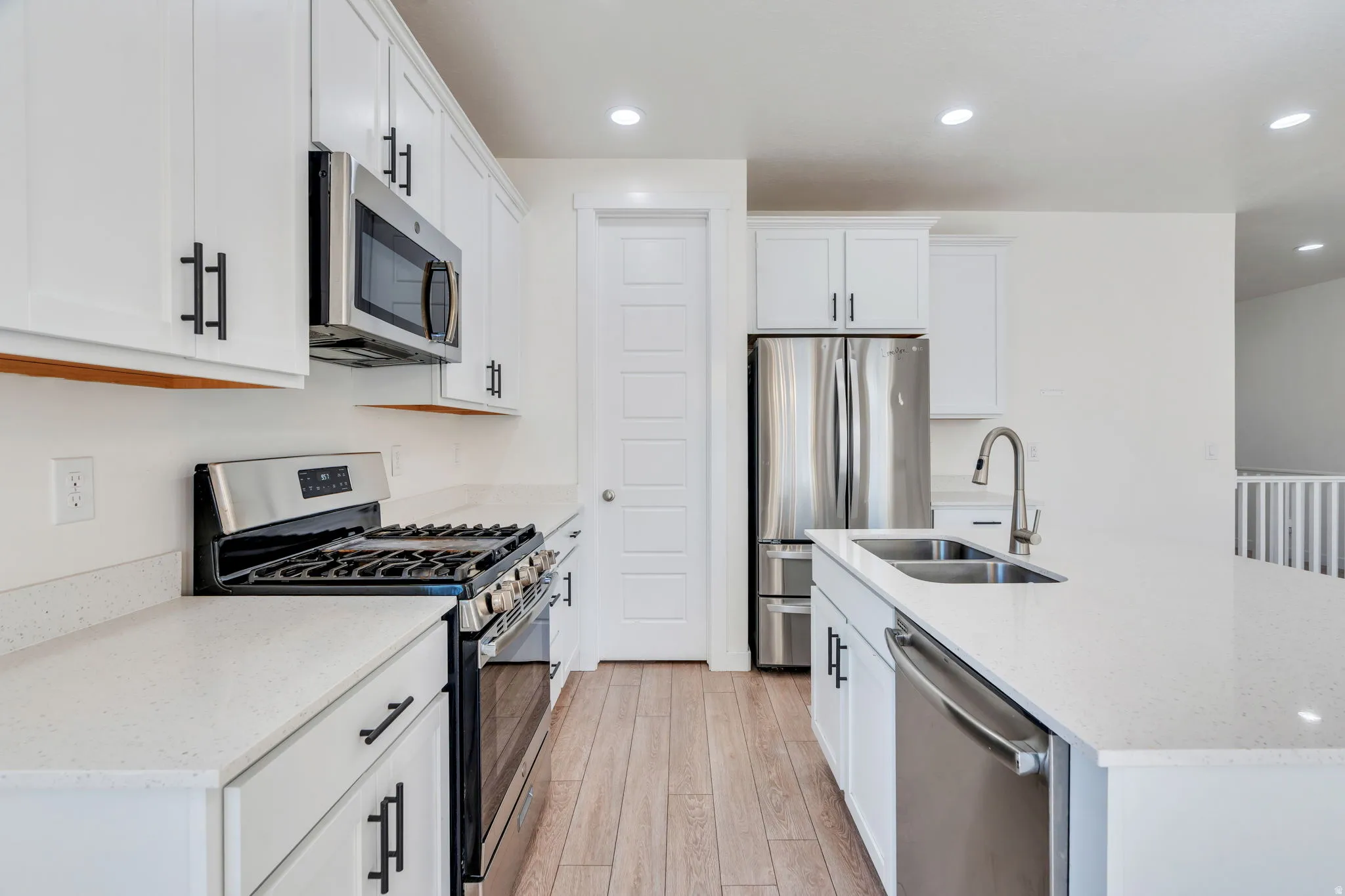 Bright kitchen featuring white cabinetry with matte black hardware, stainless steel appliances, light-toned wood-finish flooring, and white countertops