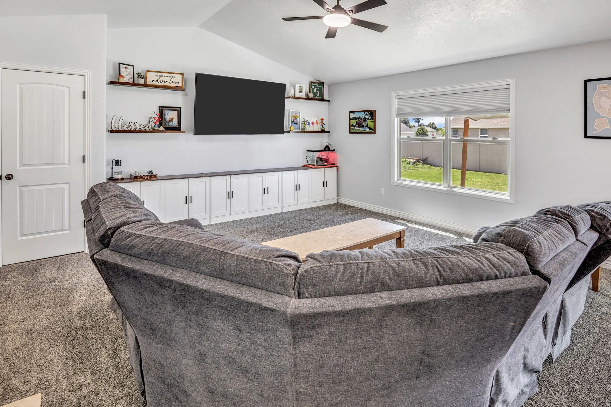 Living area featuring vaulted ceilings with a ceiling fan, a large window offering outdoor light, built-in white cabinetry with a dark countertop, and floating wall shelves