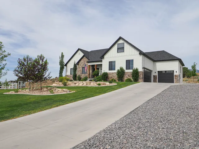 Contemporary farmhouse-style residence featuring white vertical and horizontal siding, stone accents, dark-framed windows, a dark shingle roof, and multiple dark garage doors