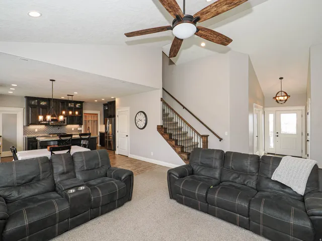 Vaulted ceiling living area with a dark wood-finish ceiling fan, staircase with wood treads and metal balusters, and a kitchen featuring dark cabinetry, stainless steel appliances, and pendant lighting