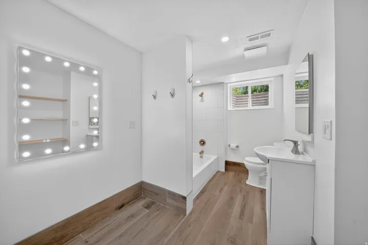 Bathroom featuring wood-finish flooring, a contemporary vanity with a white countertop, a built-in bathtub with white subway tile surround, recessed lighting, and a large illuminated wall mirror
