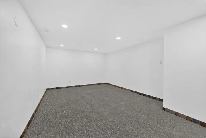 Spacious room featuring neutral carpeting, white textured walls, recessed ceiling lighting, and a dark tile baseboard