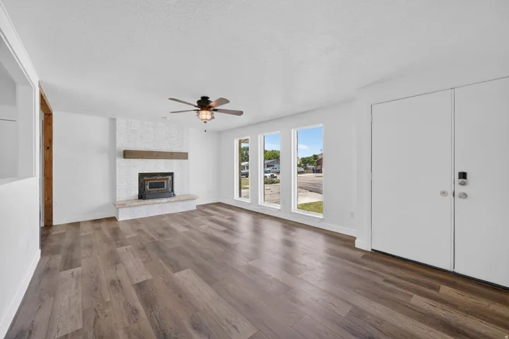 Bright living area featuring wood-finish flooring, a white textured fireplace with a stone hearth and wood mantel, three large windows, and a ceiling fan with integrated lighting
