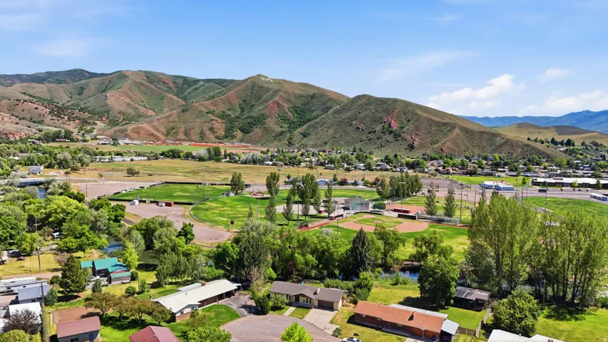 Expansive community with varied architecture and mature trees, featuring multiple recreational fields, a water feature, and distant mountainous terrain