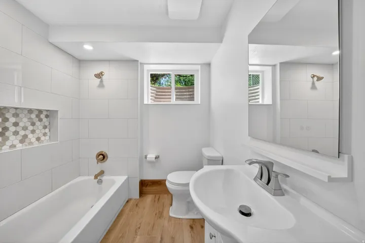 Contemporary bathroom featuring a white tile tub surround with an integrated hexagonal mosaic niche, a deep soaking tub with brushed brass fixtures, and wood-finish flooring