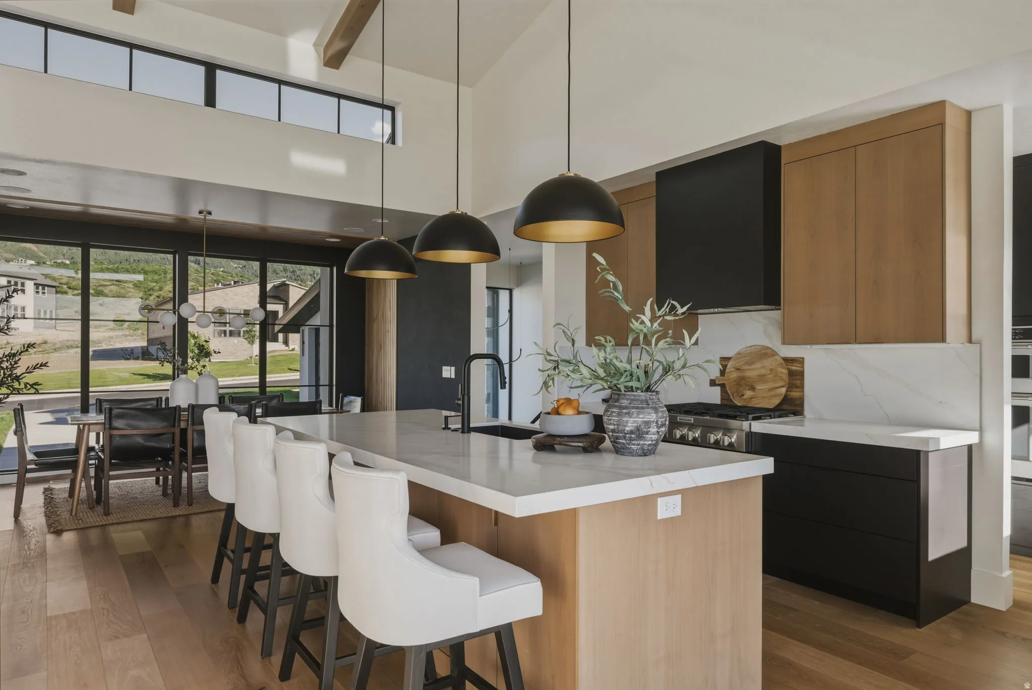 Modern kitchen featuring a large center island with integrated sink, light wood-finish cabinetry, black range hood, white countertops with matching backsplash, and exposed ceiling beams