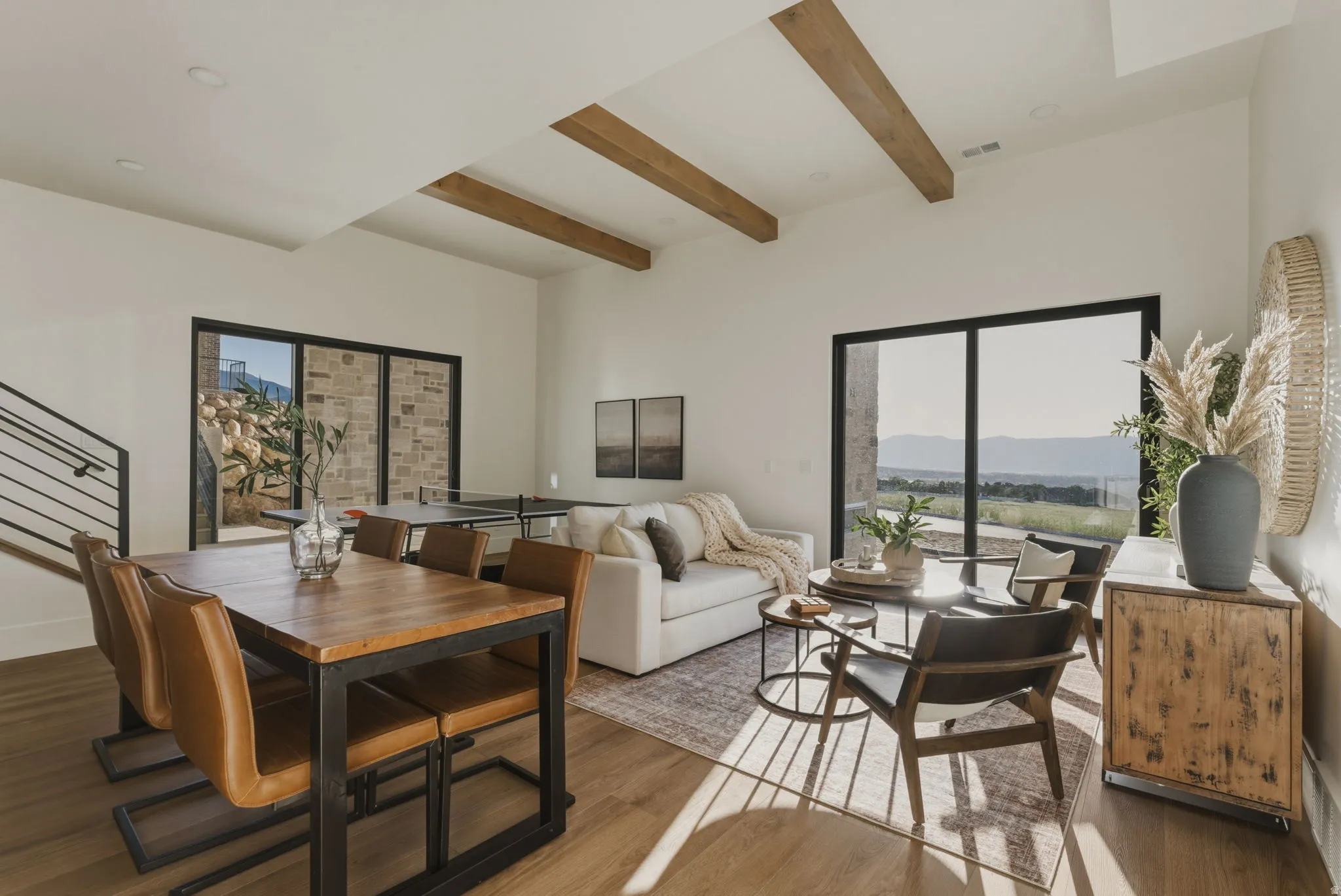 Spacious open-concept interior featuring high ceilings with exposed wood beams, expansive windows, light wood-finish flooring, and a contemporary black metal staircase railing