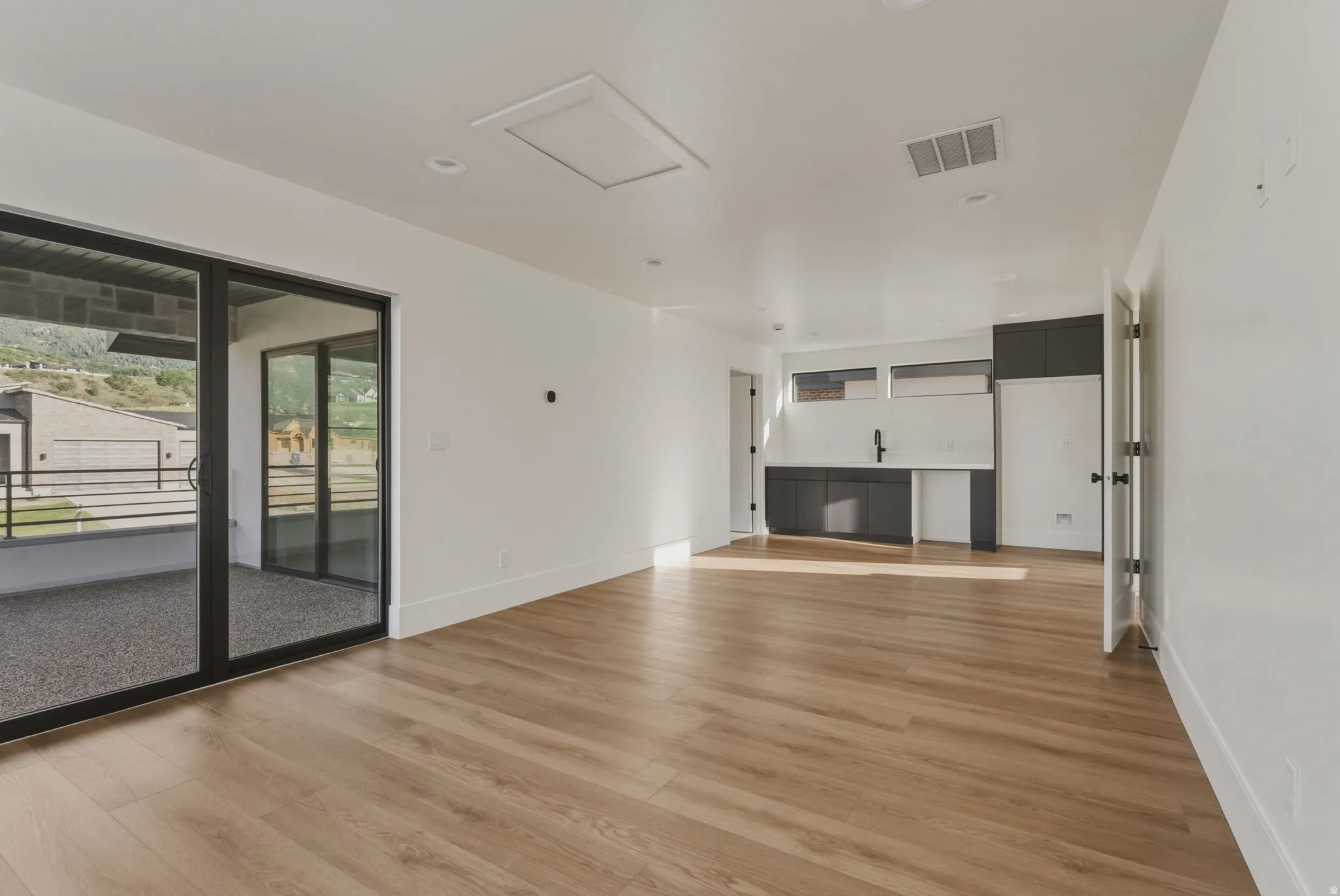 Expansive room featuring wood-finish flooring, a contemporary wet bar with dark cabinetry, and a large sliding glass door opening to an outdoor patio with a mountain backdrop